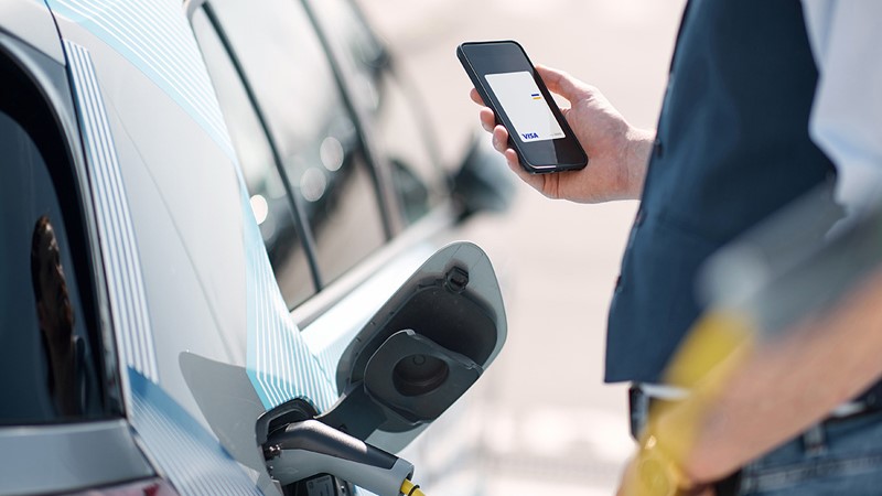 Mensch lädt sein Elektroauto und hält Smartphone mit hinterlegter Visa Karte in der Hand.