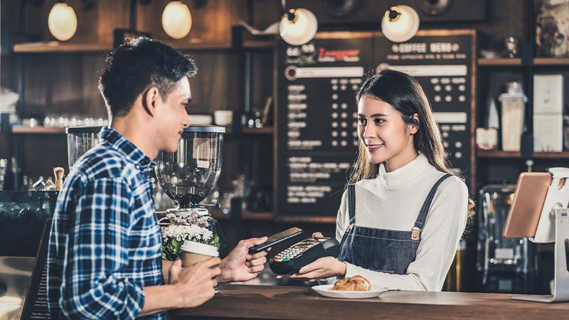 Mann bezahlt Kaffee per Smartphone bei Kassiererin, die ihm Bezahlterminal entgegenreicht.